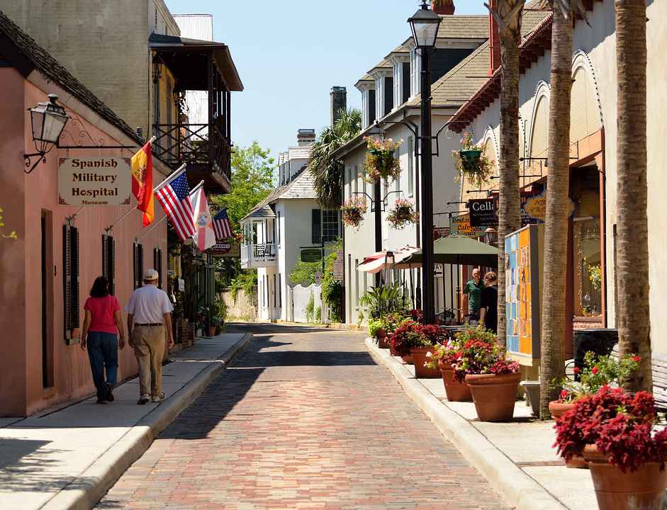 Image is outdoors and contains two people walking down a street with shops on each side.